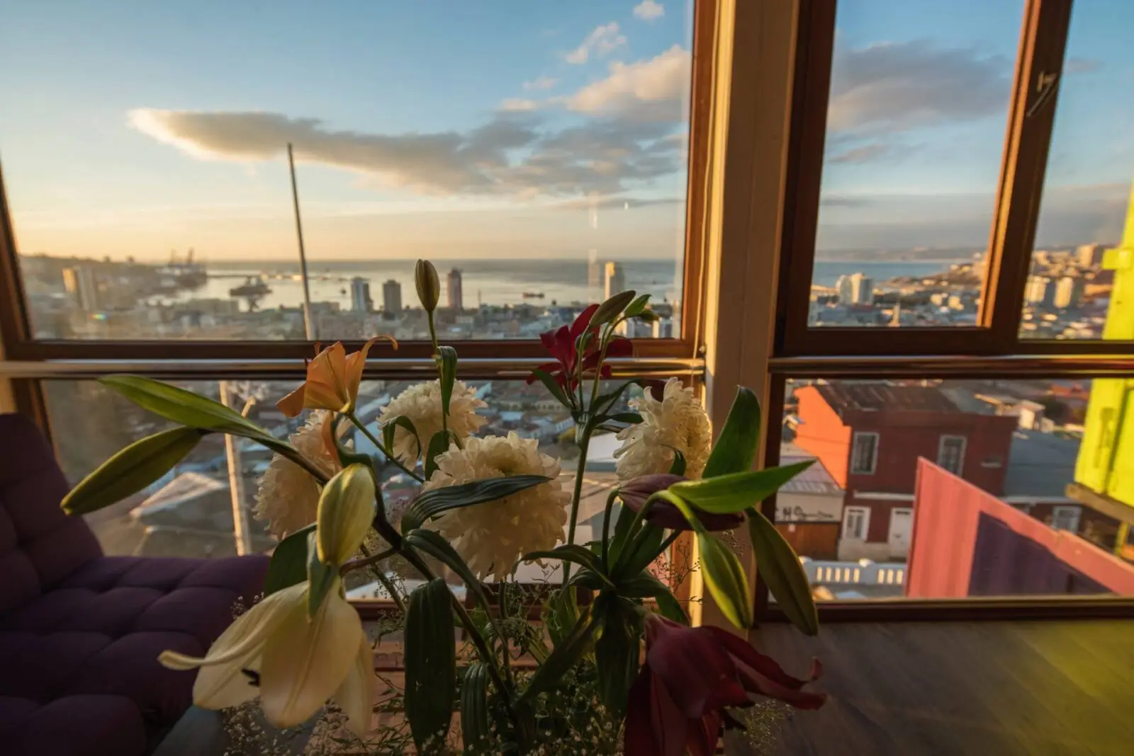 A bouquet of lilies and white flowers sits on a table by large windows, overlooking a cityscape and a harbor with ships at sunset. Warm sunlight fills the room and colorful buildings are visible outside.