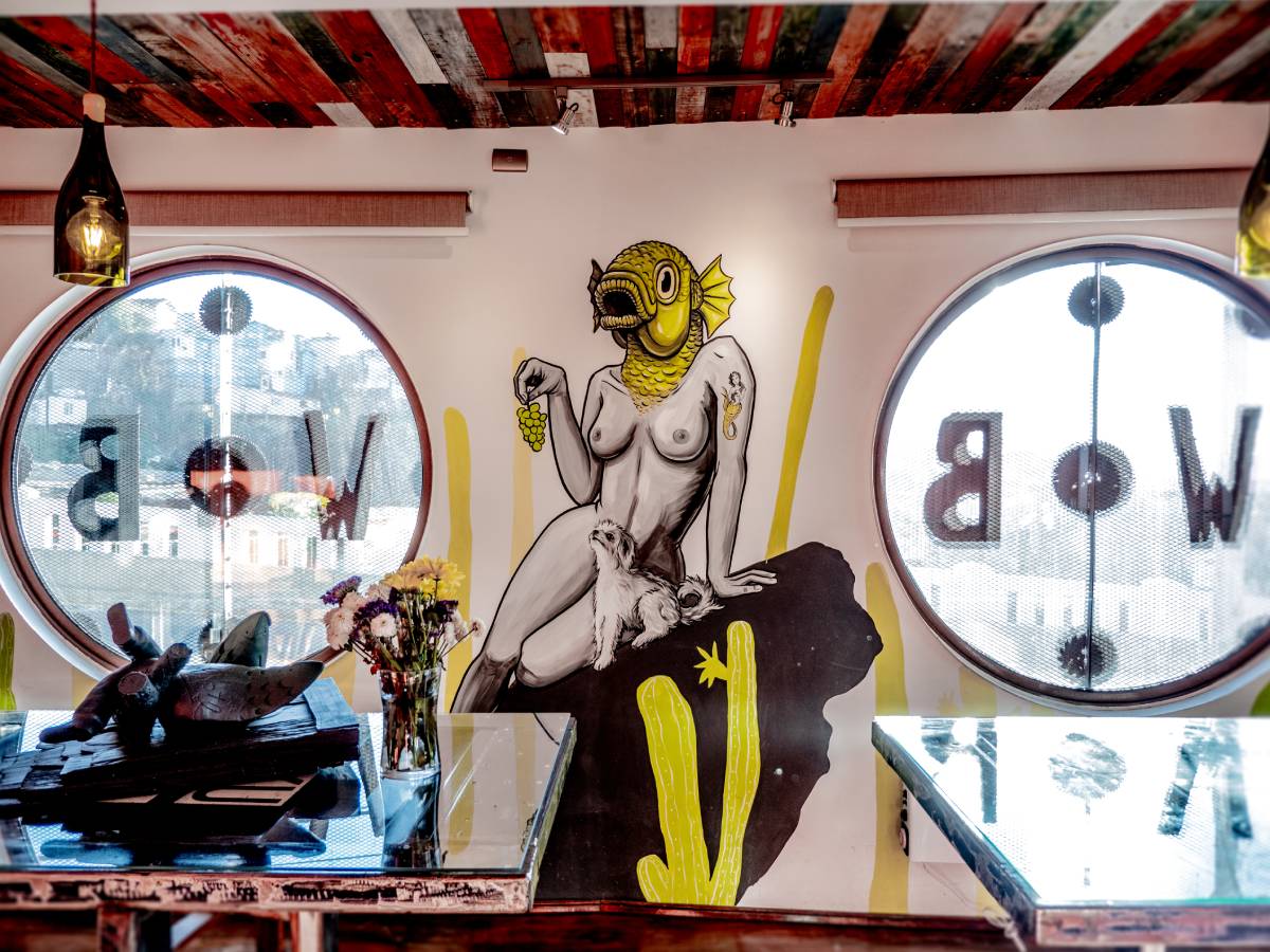 A bright room with two circular windows and vibrant wall art of a nude human figure with a yellow fish head, holding grapes. Flowers and decorative items sit on mirrored tables in the foreground.