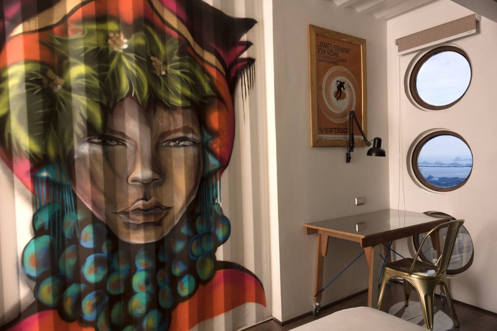 A modern room features a colorful mural of a womans face with leafy hair, a small desk with a metallic chair, a wall lamp, a poster, and two round windows overlooking a body of water.