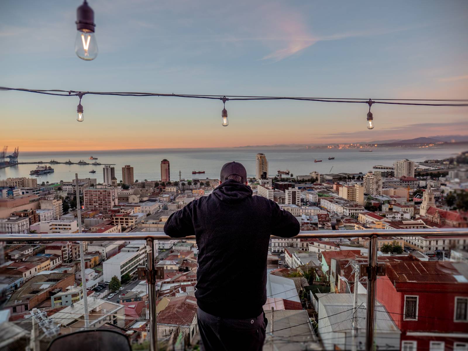 A person in a dark hoodie leans on a glass railing, overlooking a coastal city at sunset with buildings, ships in the bay, and distant hills under hanging string lights.