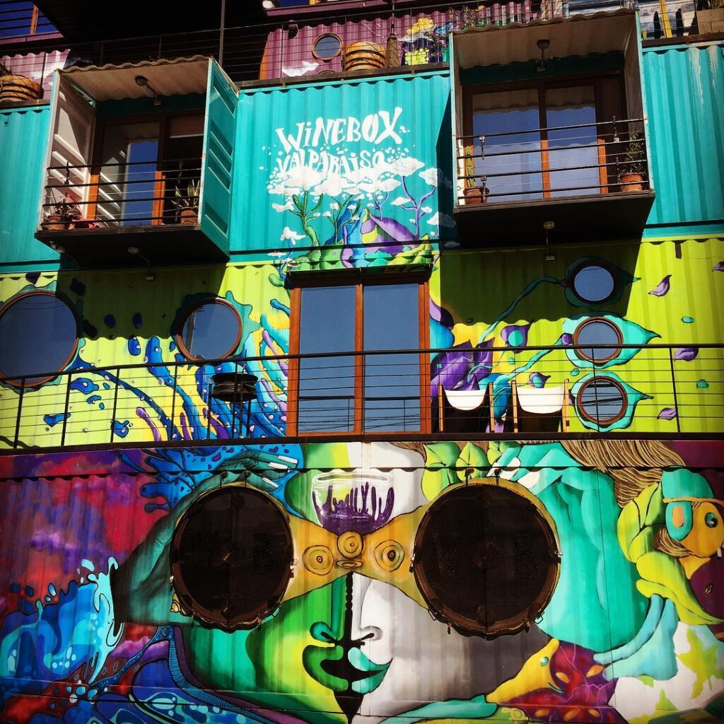Colorful shipping containers stacked as a building, with balconies and large mural art featuring vibrant abstract faces and shapes. “Winebox Valparaiso” is painted on one of the containers.