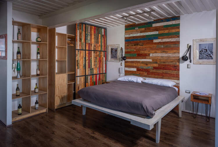 A modern bedroom with a wooden floor, a platform bed with gray bedding, a colorful reclaimed wood accent wall, open shelves with bottles, a small nightstand, and wall-mounted reading lights.