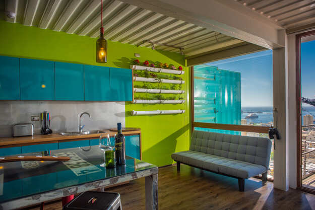 Modern, colorful apartment with blue cabinets, green walls, wall-mounted planters, a gray futon, and large windows offering a view of the sea and cityscape. Natural light fills the room.